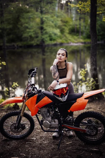 Charmig tatuerad racer flicka bär motocross outfit med halvnaken bål lutar sig på sin cykel och tittar på kameran i skogen — Stockfoto