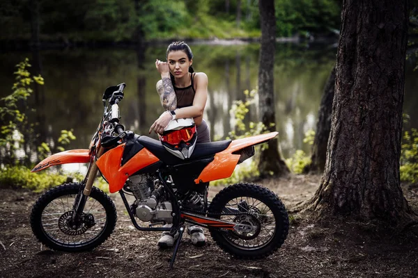 Affascinante ragazza pilota tatuata indossando abito da motocross con busto semi nudo appoggiato sulla sua moto e guardando sulla macchina fotografica nel bosco — Foto Stock