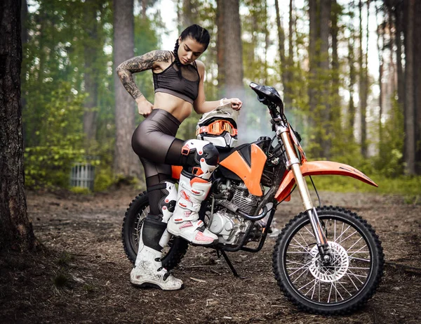 Tatuerad racer flicka bär motocross outfit med halvnaken bål poserar bredvid hennes bikein skogen — Stockfoto