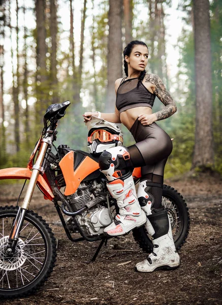 Motokros kıyafeti giyen, yarı çıplak vücutlu, ormanda bisikletinin yanında poz veren dövmeli yarışçı kız. — Stok fotoğraf