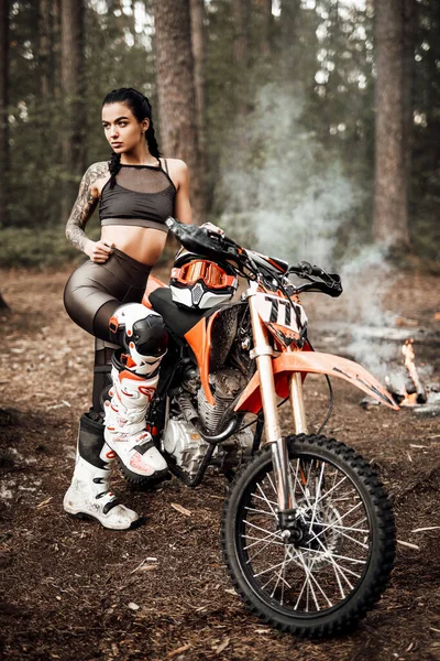 Motokros kıyafeti giyen, yarı çıplak vücutlu, ormanda bisikletinin yanında poz veren dövmeli yarışçı kız. — Stok fotoğraf