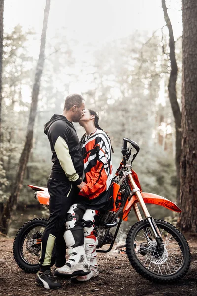 Pár csók áll egy motocross kerékpár mellett és tábortűz az erdőben — Stock Fotó