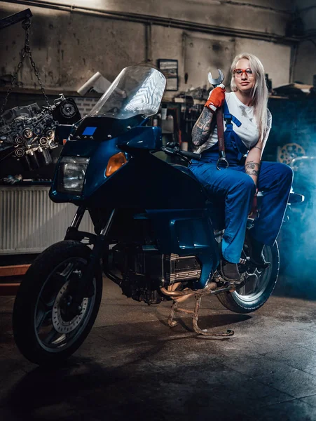 Mulher loira mecânico no trabalho macacão segurar chave grande enquanto sentado no sportbike na garagem — Fotografia de Stock