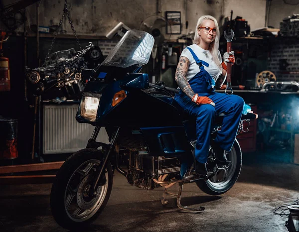 Ξανθιά γυναίκα μηχανικός στην εργασία φόρμες κατέχουν μεγάλο κλειδί, ενώ κάθεται σε sportbike στο γκαράζ — Φωτογραφία Αρχείου