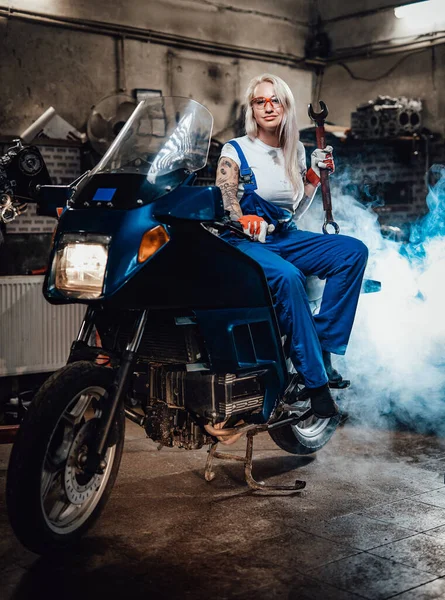 Mulher loira mecânico no trabalho macacão segurar chave grande enquanto sentado no sportbike na garagem — Fotografia de Stock