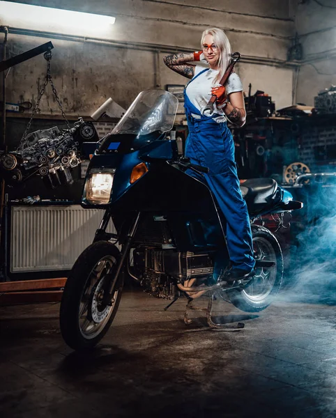 Ξανθιά γυναίκα μηχανικός με τατουάζ χέρια φορώντας φόρμες εργασίας ποζάροντας στο sportbike της στο γκαράζ — Φωτογραφία Αρχείου