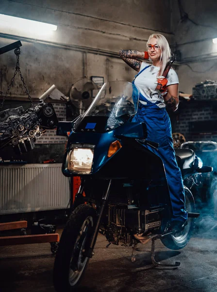 Loira mecânica feminina com as mãos tatuadas vestindo macacões de trabalho posando em seu sportbike na garagem — Fotografia de Stock