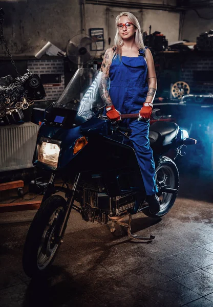 Blond kvinnlig mekaniker med tatuerade händer bär arbete overaller poserar på sin sportcykel i garage — Stockfoto