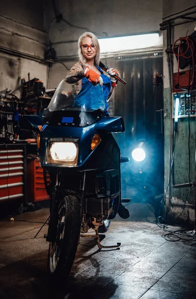 Ξανθιά γυναίκα μηχανικός με τατουάζ χέρια φορώντας φόρμες εργασίας ποζάροντας στο sportbike της στο γκαράζ — Φωτογραφία Αρχείου