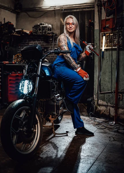 Feminino tatuado mecânico no trabalho macacão segurar uma chave grande e posando para uma câmera na garagem — Fotografia de Stock