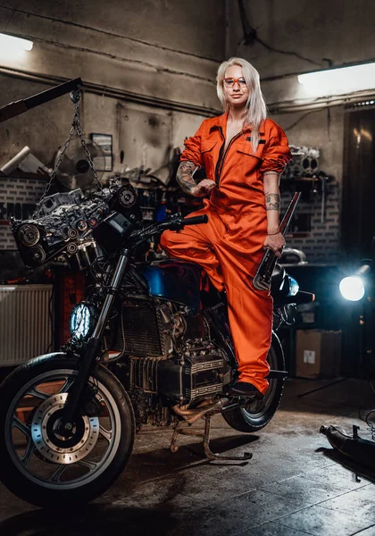 Όμορφη γυναίκα μηχανικός σε πορτοκαλί φόρμα κρατήσει ένα μεγάλο κλειδί και ποζάρουν για μια φωτογραφική μηχανή στο γκαράζ — Φωτογραφία Αρχείου