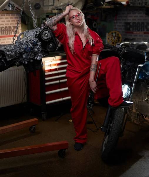 Blond female mechanic with tattooed hands in orange overalls stands in garage or workshop