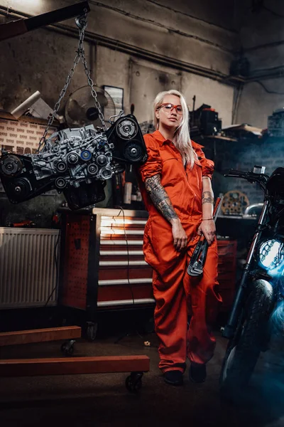 Mecânico fêmea loira com as mãos tatuadas em macacão laranja fica na garagem ou oficina — Fotografia de Stock