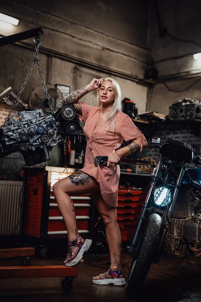 Vacker blond flicka med tatuerad kropp bär rosa klänning poserar i garage eller verkstad — Stockfoto