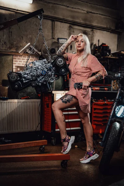 Bella ragazza bionda con il corpo tatuato che indossa un abito rosa in posa in garage o in officina — Foto Stock