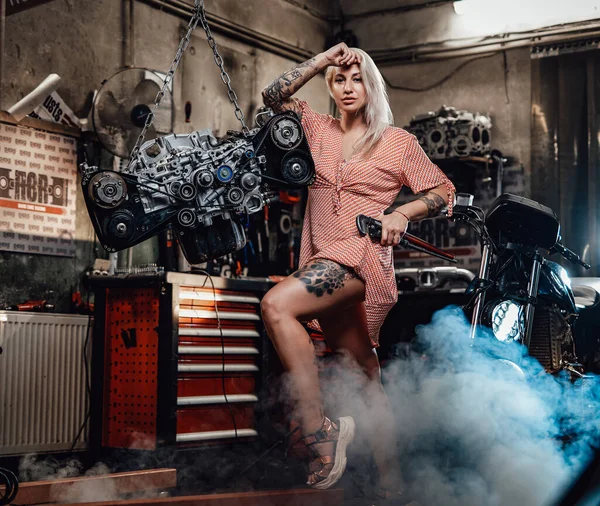 Belle fille blonde avec un corps tatoué portant une robe rose posant dans un garage ou un atelier — Photo