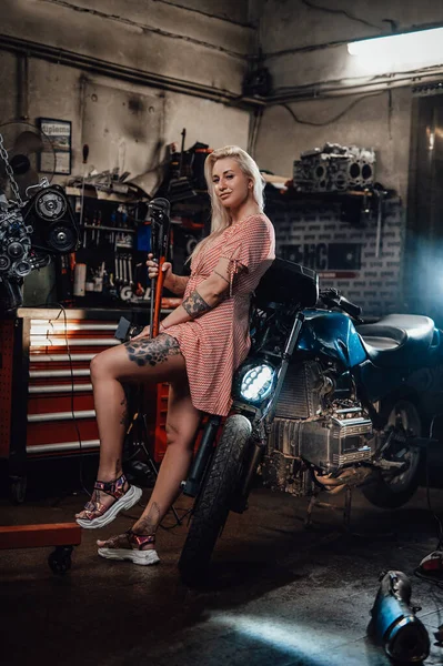 Menina loira bonita com corpo tatuado vestindo vestido rosa posando na garagem ou oficina — Fotografia de Stock