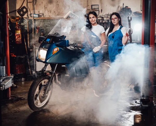 Δύο γυναίκες μηχανικός ποζάρουν δίπλα σε ένα sportbike στο αυθεντικό γκαράζ εργαστήριο που περιβάλλεται από καπνό — Φωτογραφία Αρχείου