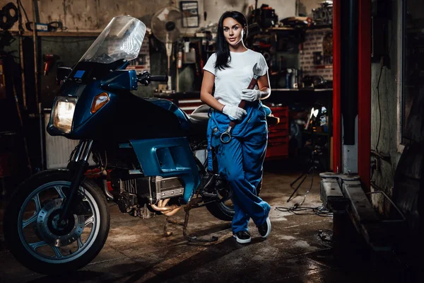 Mulher morena bonita em macacão azul posando ao lado de um sportbike na garagem ou oficina — Fotografia de Stock