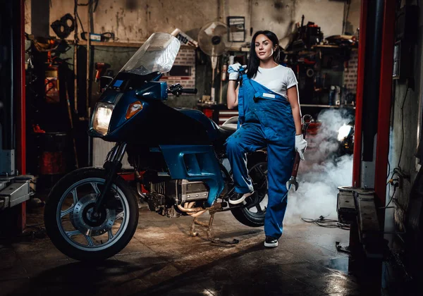 Mulher morena bonita em macacão azul posando ao lado de um sportbike na garagem ou oficina — Fotografia de Stock