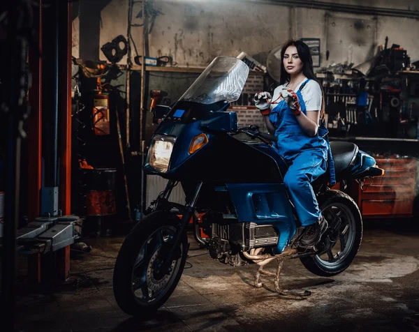 Jovem morena mecânica feminina em macacões azuis sentada em sportbike na garagem ou oficina — Fotografia de Stock