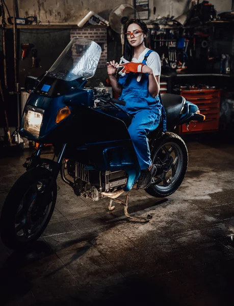 Jovem morena mecânica feminina em macacões azuis sentada em sportbike na garagem ou oficina — Fotografia de Stock
