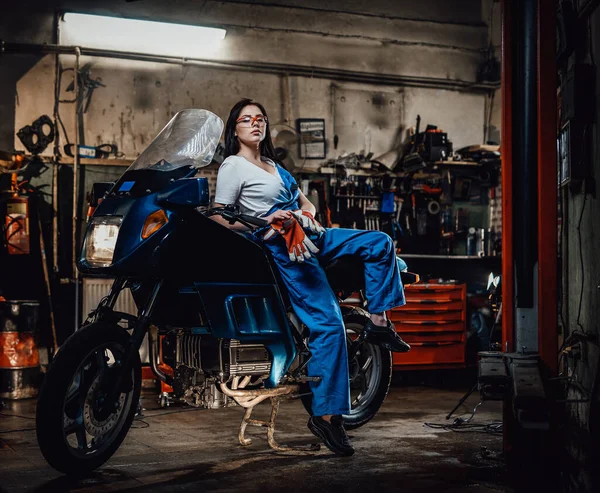 在车库里或车间里，穿着蓝色工作服的性感黑发女郎摆出姿势，摆出相机的姿势，靠在摩托车上 — 图库照片
