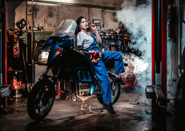 Menina morena quente em macacão azul posando para uma câmera enquanto se inclina no sportbike na garagem ou oficina — Fotografia de Stock