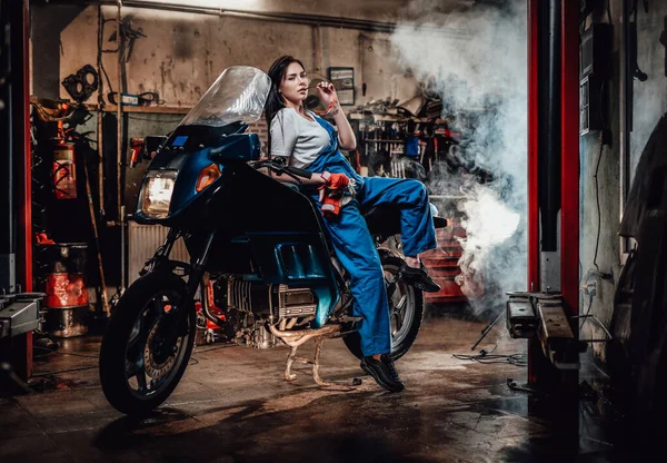 Chica morena caliente en mono azul posando para una cámara mientras se apoya en la bicicleta deportiva en el garaje o taller — Foto de Stock