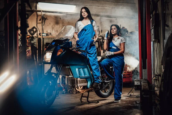 Duas mulheres morenas de macacão azul posando ao lado de um sportbike na garagem oficina autêntica — Fotografia de Stock