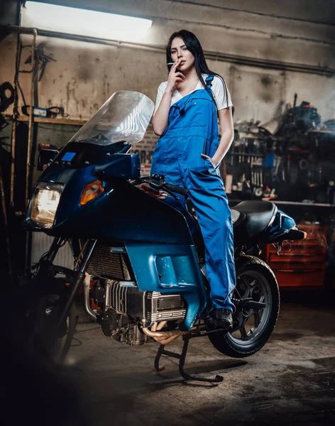 Γυναίκα μηχανικός χαλαρωτικό κάπνισμα ένα τσιγάρο, ενώ στέκεται σε sportbike στο γκαράζ ή εργαστήριο — Φωτογραφία Αρχείου