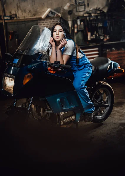 Belle brune mécanicienne fumant une cigarette tout en étant assise sur un vélo de sport dans un garage — Photo