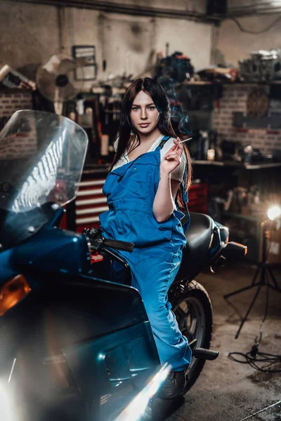 ガレージでスポーツバイクに座っている間にタバコを吸う美しいブルネットの女性整備士 — ストック写真