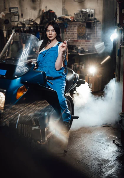 Linda morena mecânica feminina fumar um cigarro enquanto sentado em sportbike na garagem — Fotografia de Stock
