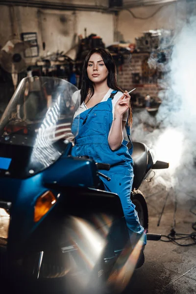 Güzel esmer kadın tamirci garajda spor bisikleti üzerinde otururken sigara içiyor. — Stok fotoğraf