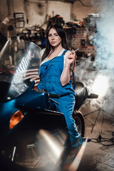 Hermosa morena motorista o mecánico en mono azul relajante fumar un cigarrillo mientras está sentado en moto deportiva en el garaje o taller — Foto de Stock