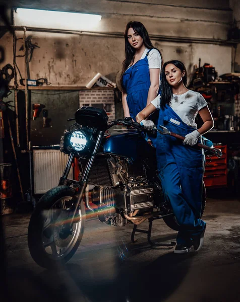 Δύο μελαχρινές γυναίκες με μπλε φόρμα ποζάρουν δίπλα σε έναν ειδικό προφυλακτήρα στο αυθεντικό γκαράζ του εργαστηρίου. — Φωτογραφία Αρχείου