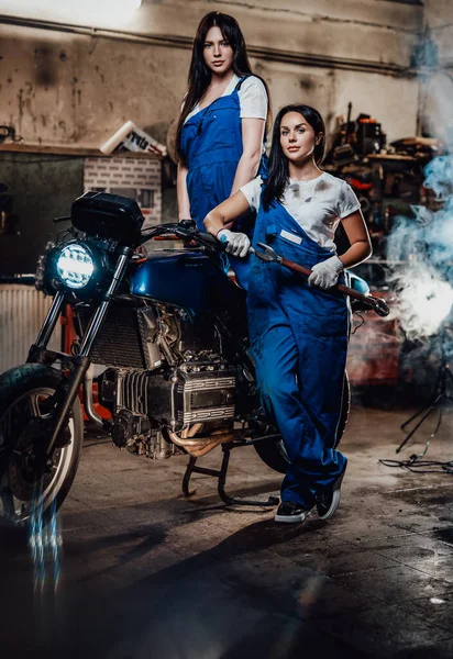 Duas mulheres morenas de macacão azul posando ao lado de um bobber personalizado na garagem oficina autêntica — Fotografia de Stock