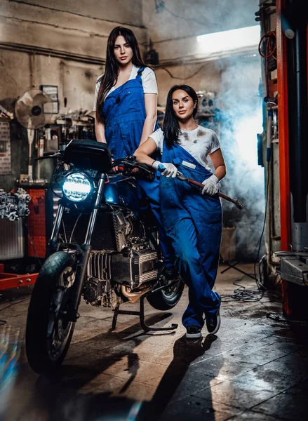 Duas mulheres morenas de macacão azul posando ao lado de um bobber personalizado na garagem oficina autêntica — Fotografia de Stock