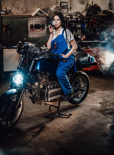 Mooie brunette vrouwelijke monteur roken een sigaret zitten op aangepaste bobber in garage — Stockfoto