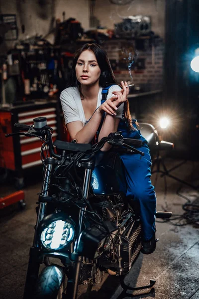 Linda morena motociclista ou mecânico relaxante fumar um cigarro na garagem ou oficina — Fotografia de Stock