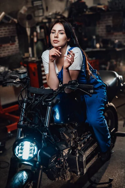 Linda morena motociclista ou mecânico relaxante fumar um cigarro na garagem ou oficina — Fotografia de Stock