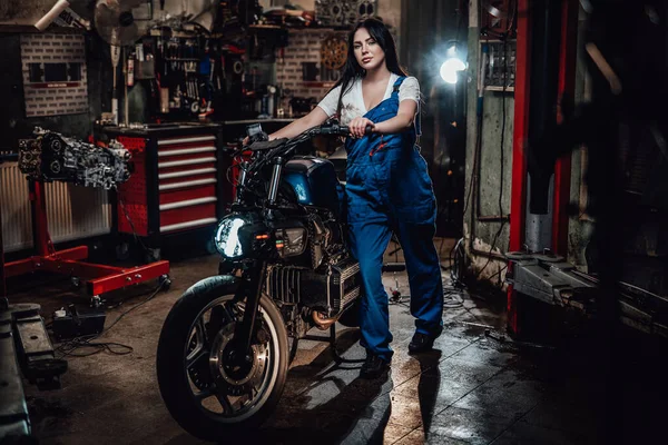 Linda jovem mecânica feminina em macacão azul posando com bobber personalizado na garagem ou oficina — Fotografia de Stock