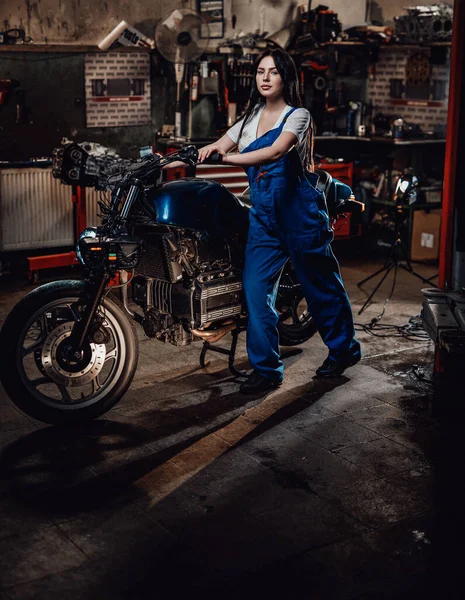 Όμορφη νεαρή γυναίκα μηχανικός σε μπλε φόρμες ποζάρουν με έθιμο bobber στο γκαράζ ή εργαστήριο — Φωτογραφία Αρχείου