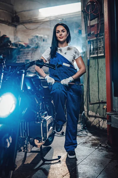 Mulher morena bonita em macacão azul posando ao lado de um bobber personalizado na garagem ou oficina — Fotografia de Stock