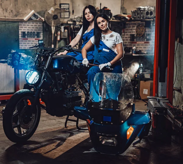 Duas mulheres morenas de macacão azul posando ao lado de um bobber personalizado na garagem oficina autêntica — Fotografia de Stock