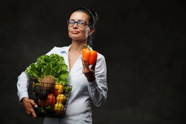 Orta yaşlı gözlüklü ve beyaz gömlekli bir kadın elinde taze sebze ve meyve sepeti tutuyor.