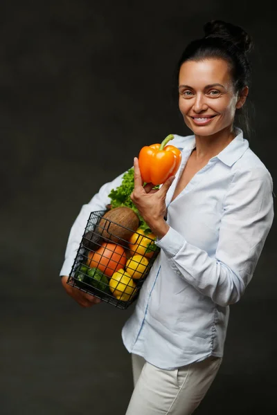 Orta yaşlı bir kadın elinde bir sepet taze sebze ve meyve tutuyor, gülümsüyor ve kameraya bakıyor. — Stok fotoğraf