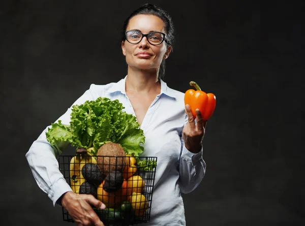 Orta yaşlı gözlüklü ve beyaz gömlekli bir kadın elinde taze sebze ve meyve sepeti tutuyor. — Stok fotoğraf