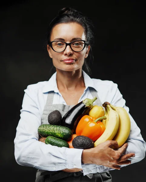앞치마를 입은 아름다운 중년의 여성 농부는 신선 한 야채와 과일을 많이 가지고 있다 — 스톡 사진